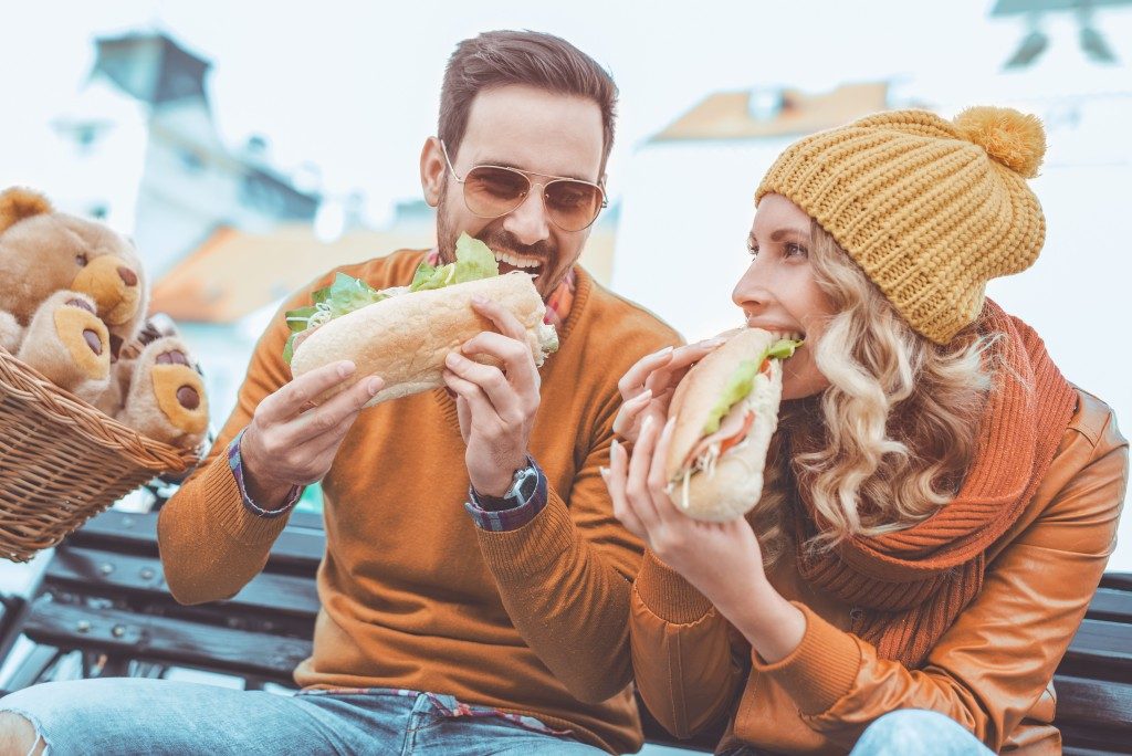 couple eating big sandwiches on a bench
